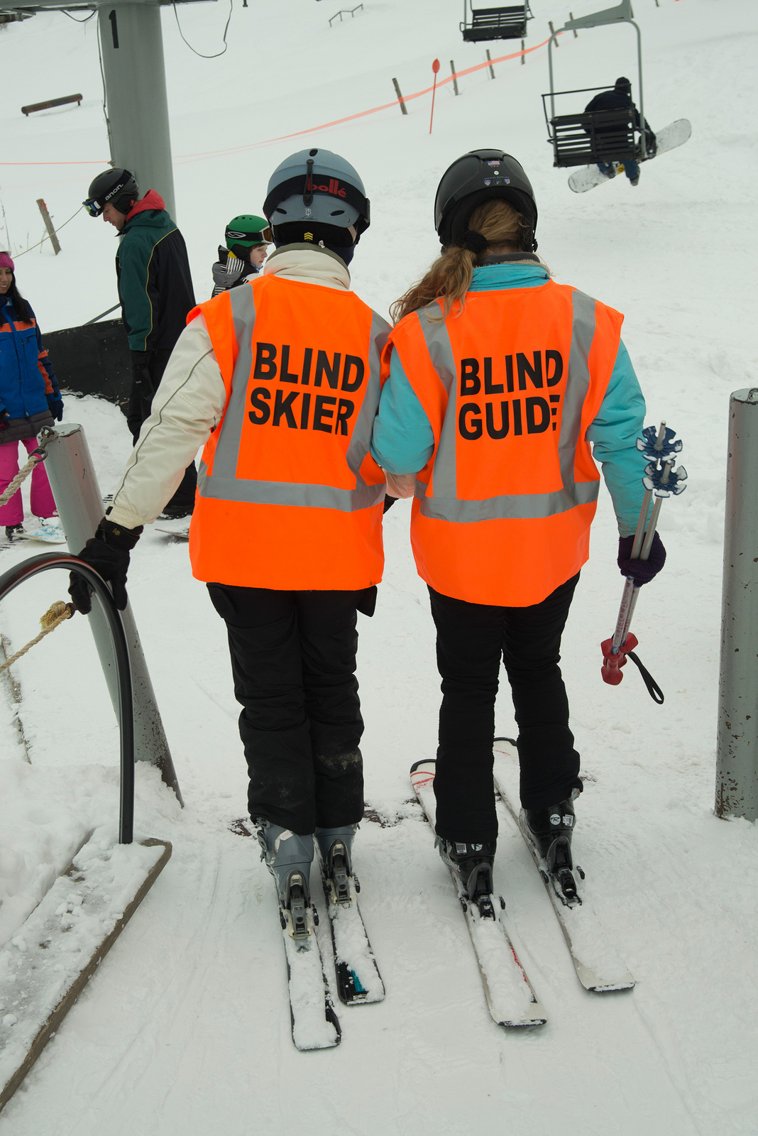 Blind guide helping blind skier onto ski lifts