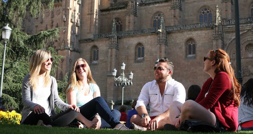 Four students sit together on the lawn in front of a large, Gothic style building